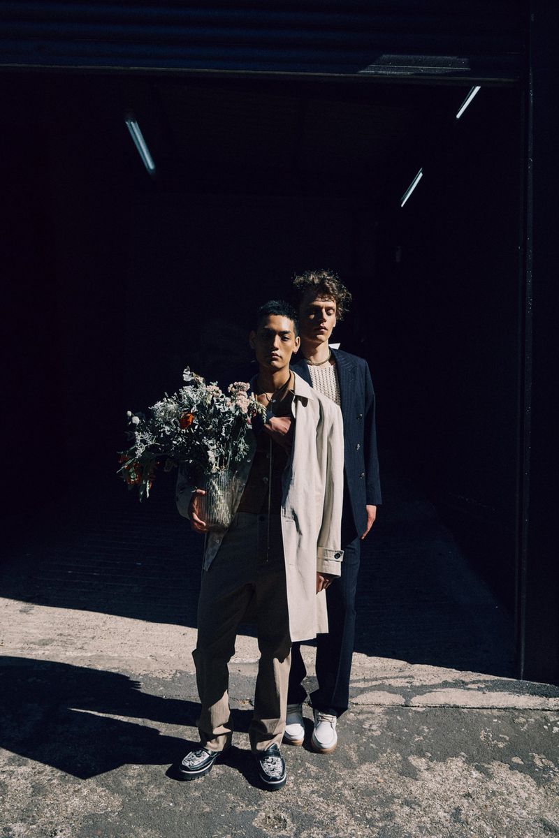 Two people in a garage-like setting. One holds a bouquet. They're wearing fashionable outerwear and shoes, bathed in bright sunlight.