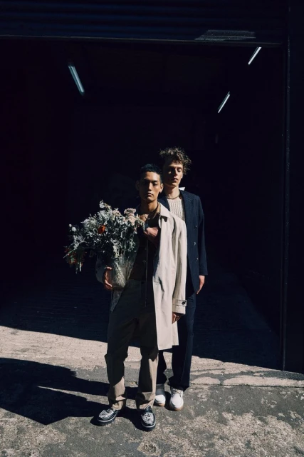 Two people in a garage-like setting. One holds a bouquet. They're wearing fashionable outerwear and shoes, bathed in bright sunlight.