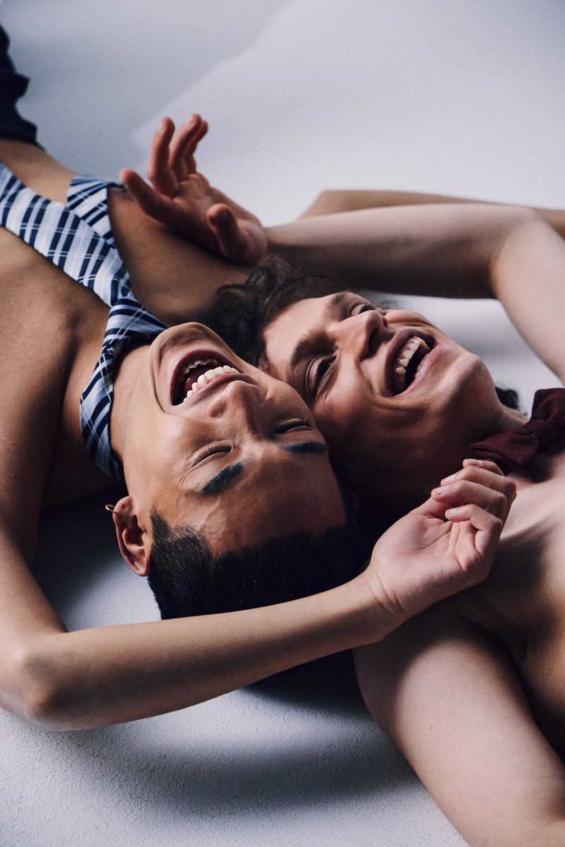 Two people laugh while lying down together, wearing ties around their necks, on a light grey background in a medium shot.
