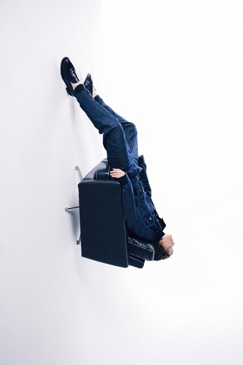 Person lying upside down on a black couch with legs and feet propped against a white wall in a brightly lit room. 