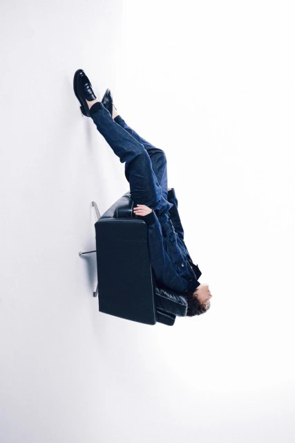 Person lying upside down on a black couch with legs and feet propped against a white wall in a brightly lit room. 