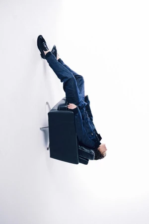 Person lying upside down on a black couch with legs and feet propped against a white wall in a brightly lit room. 