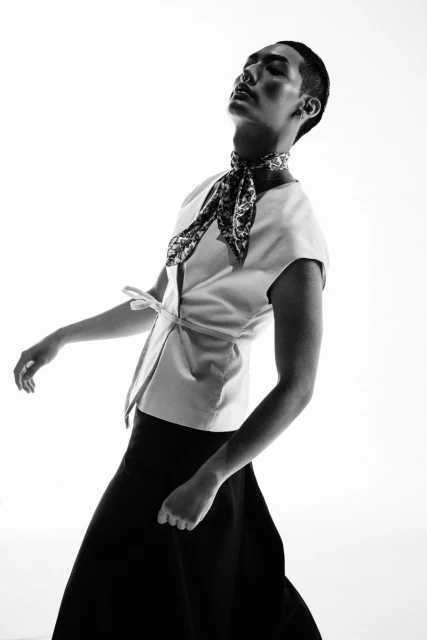 Monochrome studio portrait of a person wearing a light-colored shirt, dark skirt, and patterned neck scarf, eyes closed, head tilted back.