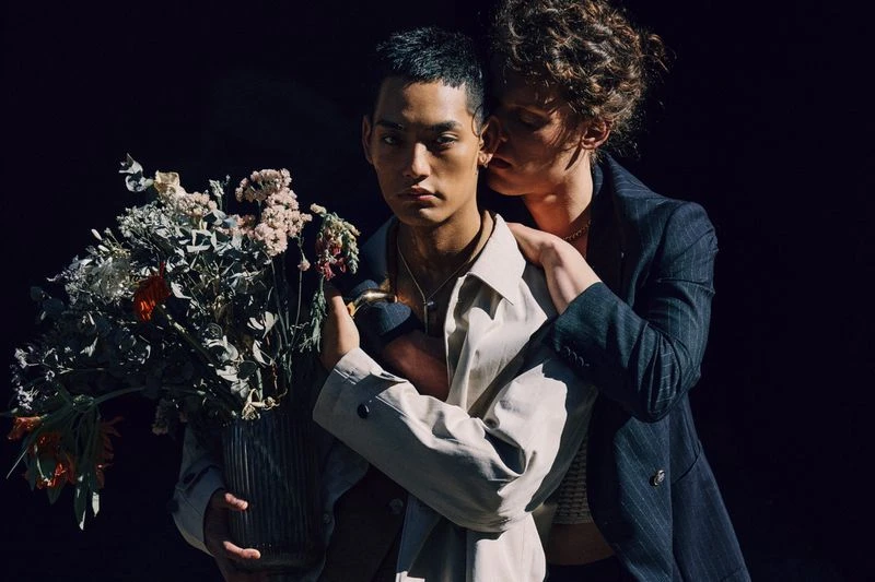Two people embrace beside a vase of flowers in a dimly lit room. One person wears a beige trench coat, the other a striped suit.