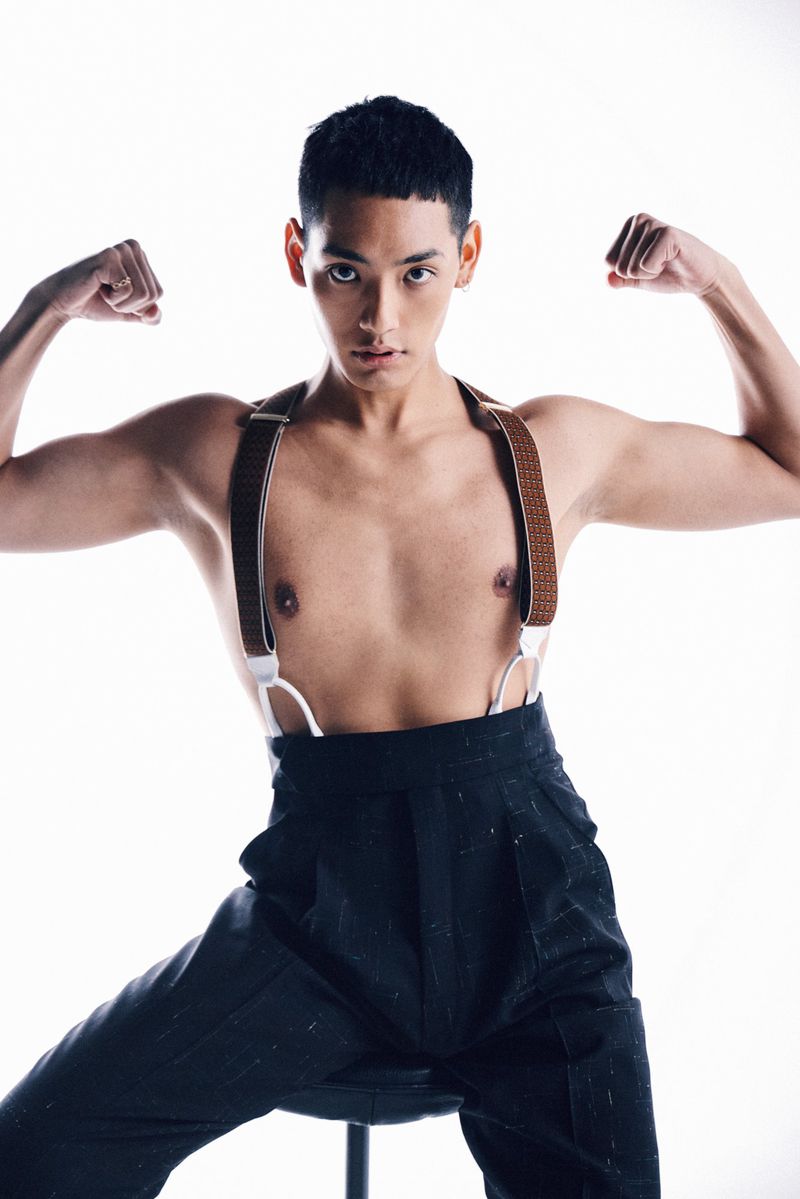 Young adult man flexing his arms while sitting on a stool, wearing suspenders and trousers in a studio setting with bright white background.