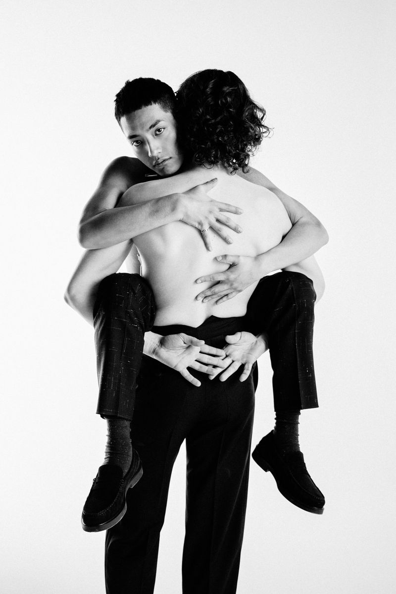 Black and white portrait of an adult male giving another adult male a piggyback ride in a bright photo studio against a white backdrop.