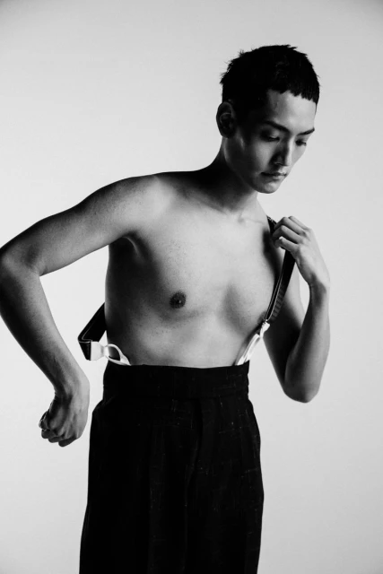 Monochrome portrait of an Asian man in black pants and suspenders, posing against a white backdrop in a photo studio with soft lighting.