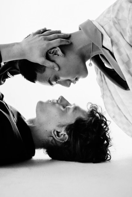 Black and white portrait of a couple face to face, one lying down and the other leaning over with hands framing their partner's face.