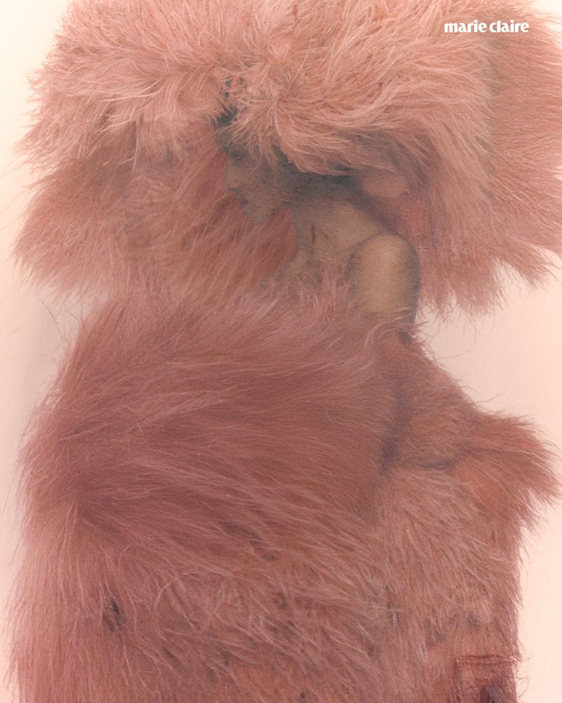 A person with light skin is partially obscured by a large, fluffy pink hat and wrap, photographed in a soft, diffused lighting.