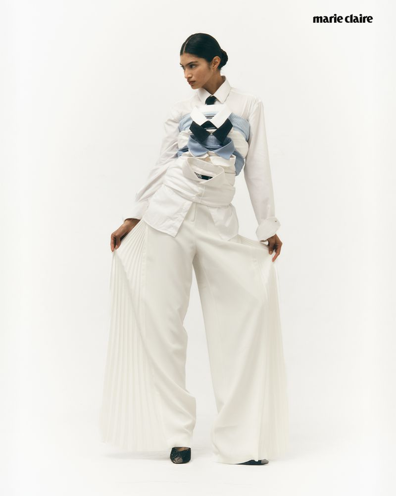 Fashion editorial shot of a woman wearing a white shirt and wide-legged pants, styled with stacked shirts and a necktie in a studio.