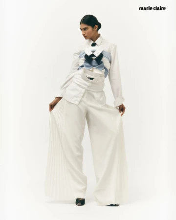 Fashion editorial shot of a woman wearing a white shirt and wide-legged pants, styled with stacked shirts and a necktie in a studio.