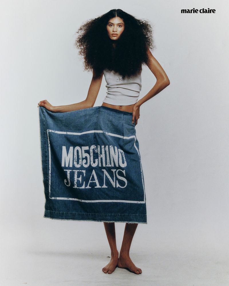 Fashion shot of a woman with afro hairstyle wearing a tank top and jeans skirt, posing in a studio with bright, neutral lighting.