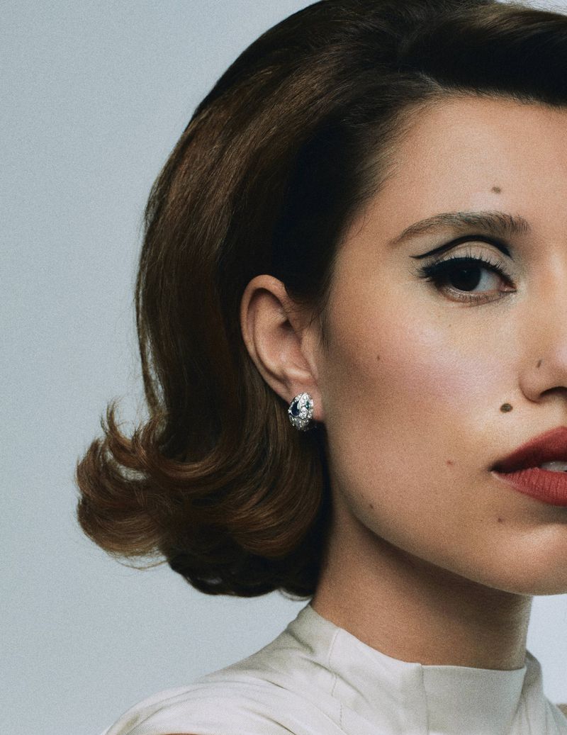 Close-up portrait of a woman with retro hairstyle and bold eye makeup, wearing a diamond earring, against a plain background.