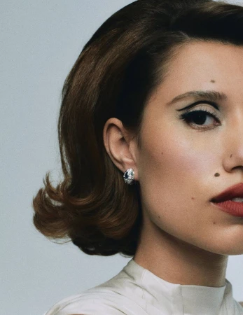 Close-up portrait of a woman with retro hairstyle and bold eye makeup, wearing a diamond earring, against a plain background.