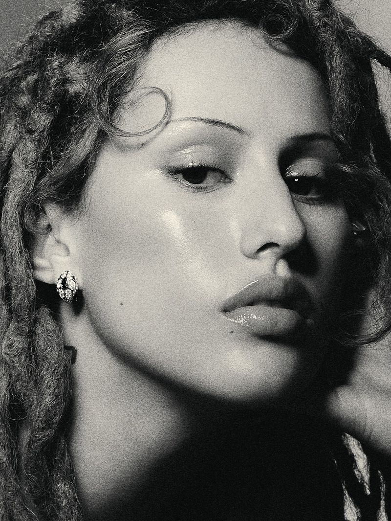 Close-up portrait of a woman with dreadlocks and a serious expression, wearing stud earrings, in a studio with shadows.