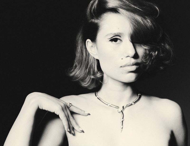 Black-and-white portrait of a young woman wearing a snake necklace and a ring, with dramatic eye makeup and styled short hair.