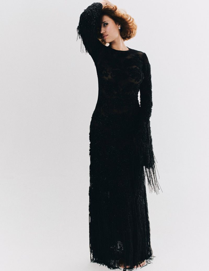 Woman in a long-sleeved black fringe dress, stands with hand on head, posing in front of white backdrop. Fashion editorial photography.