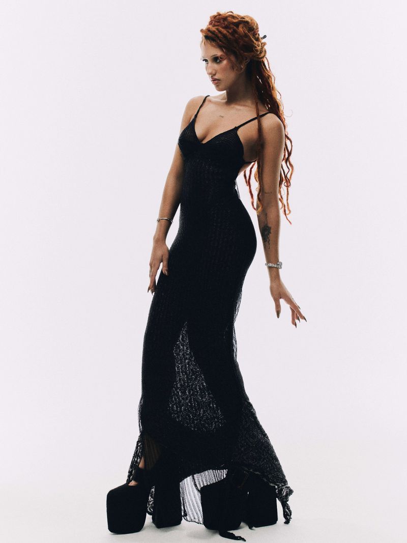 A tall, fair-skinned woman with red hair in a black dress poses in a photo studio. The model wears towering black platform heels.