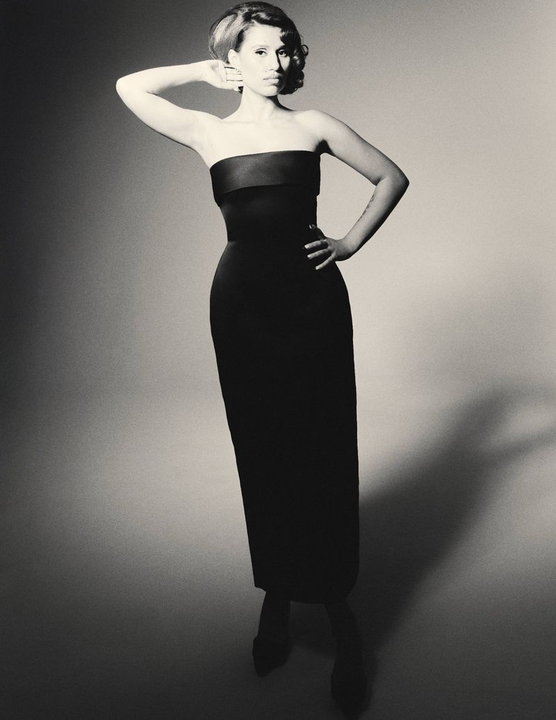 A woman in a strapless black gown poses in a studio with one hand on her head and the other on her hip.