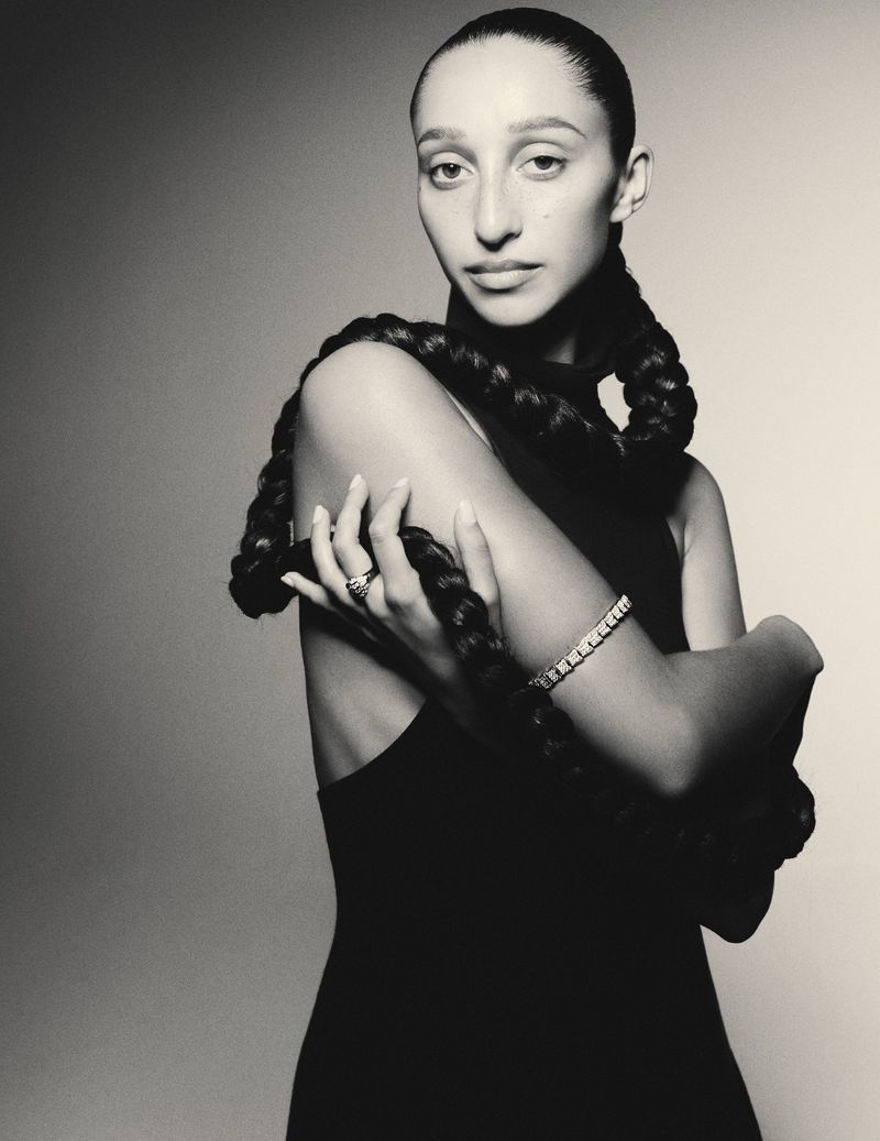 Portrait of an adult female with long dark braided hair in a black dress, wearing jewelry, posing in a photo studio against a grey background.