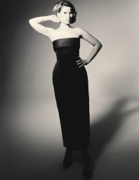 Singer Raye posing in a strapless black column gown with retro styled hair against a seamless studio backdrop.
