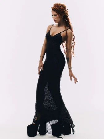 Raye posing in a sheer black maxi dress and exaggerated black platform shoes against a white studio background.