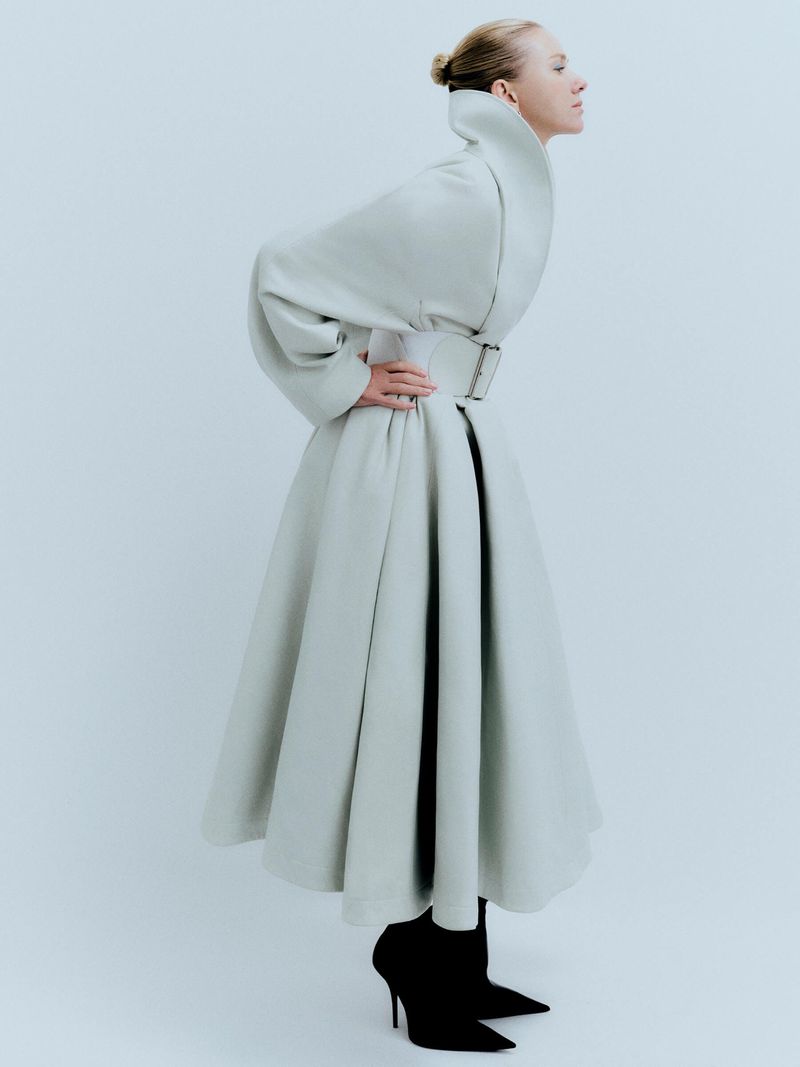 A woman in a light green dress with a high collar and belt poses in a studio with black heels. Fashion editorial style portrait.