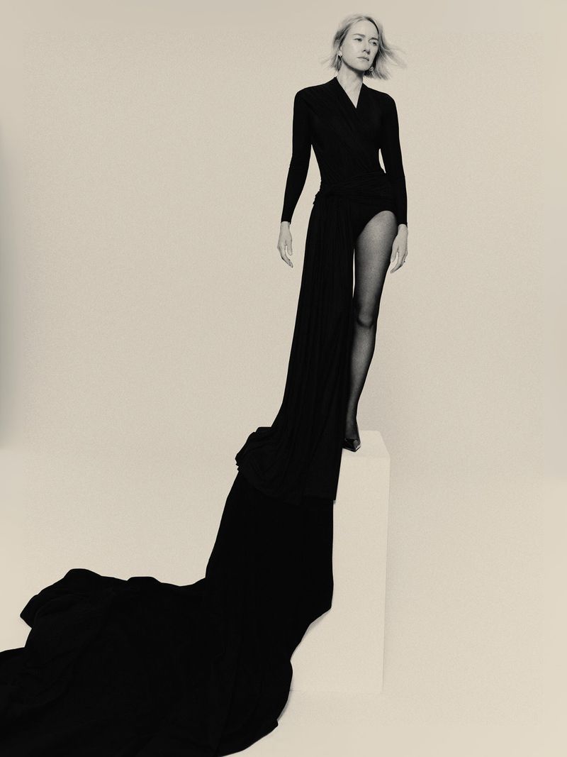 Blonde model poses in black dress and sheer black tights against white backdrop, creating high-contrast fashion studio portrait.