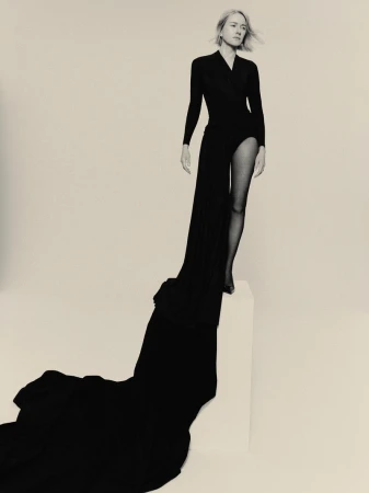 Blonde model poses in black dress and sheer black tights against white backdrop, creating high-contrast fashion studio portrait.