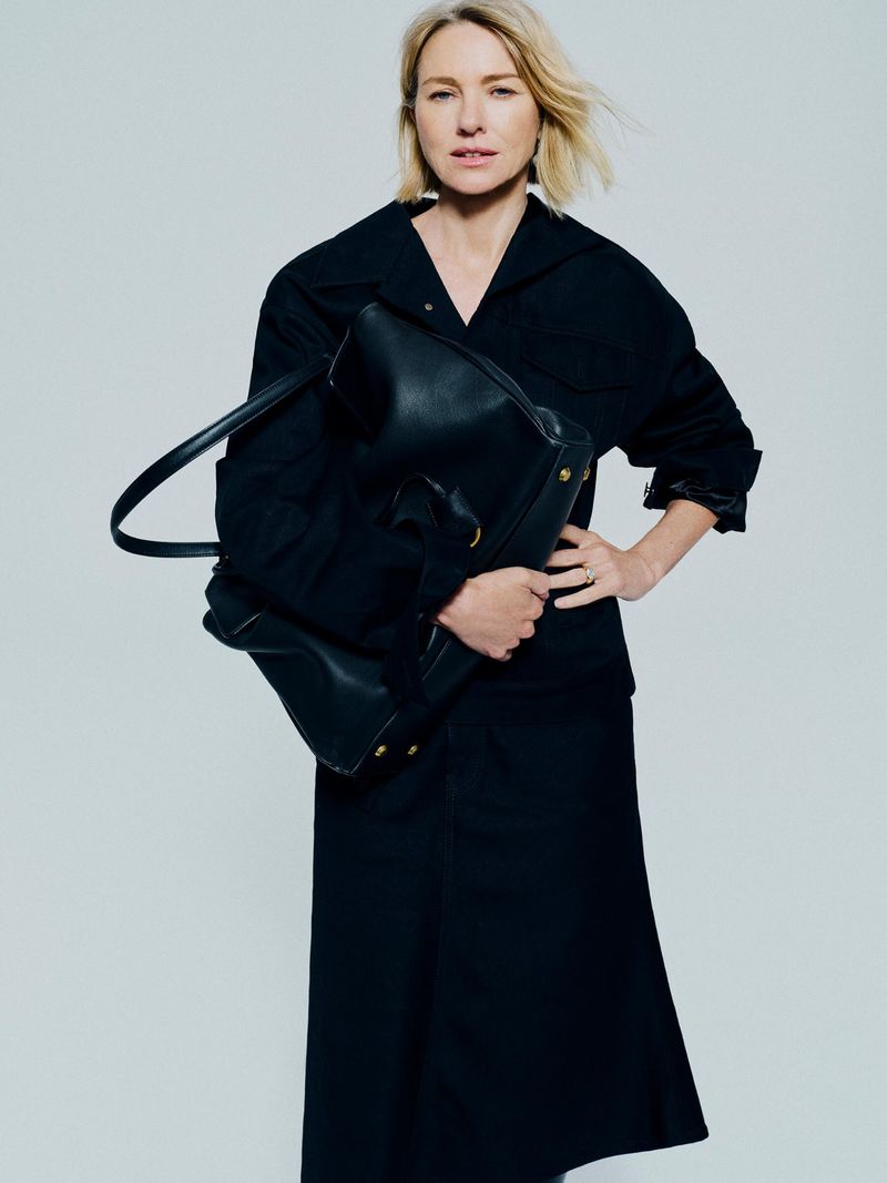 Blonde woman in a black midi dress holding a large black leather tote bag, posing in a photo studio with a neutral background.