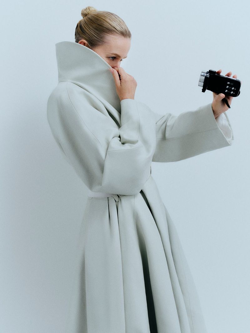 Woman in an elegant off-white coat stands in a photo studio, holding a small black video camera, posing in a fashion editorial style.