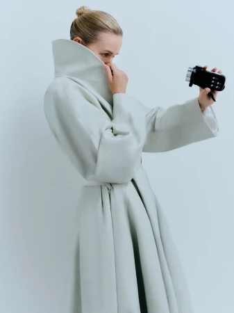 Woman in an elegant off-white coat stands in a photo studio, holding a small black video camera, posing in a fashion editorial style.