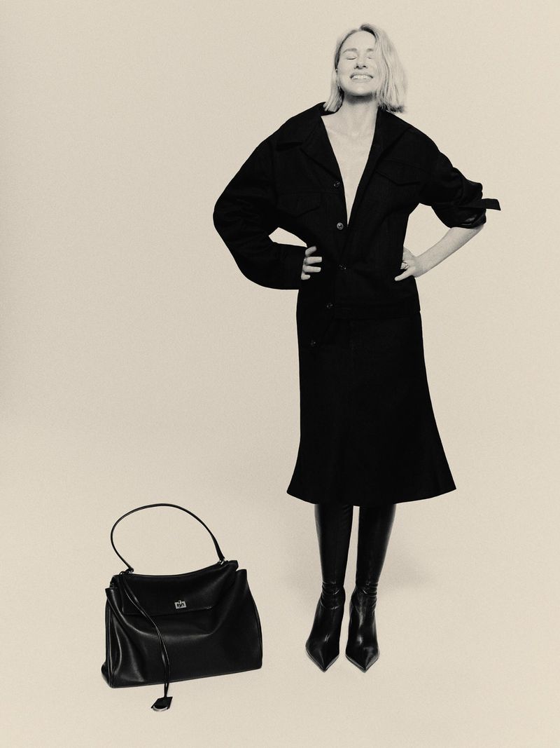 Black and white studio shot of a mature blonde woman in dark midi skirt suit with a large handbag, smiling and posing.