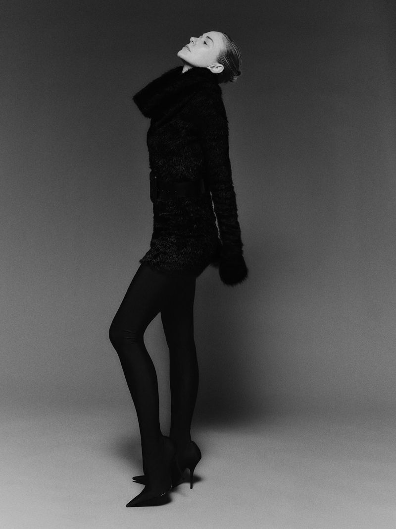 Full-length shot of a woman posing in black tights and a dark jacket with a fur collar against a solid gray backdrop in a studio.