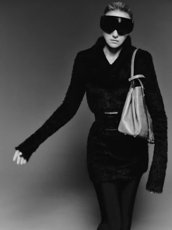 A woman in a black dress and futuristic visor poses with a handbag in a studio, creating a high-fashion, monochrome portrait.