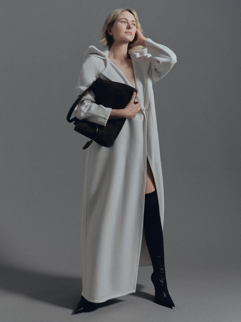 A blonde woman poses in a full-length white coat with black thigh-high boots and a black purse against a neutral gray backdrop.