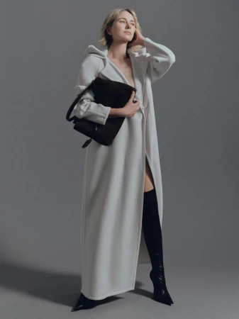 A blonde woman poses in a full-length white coat with black thigh-high boots and a black purse against a neutral gray backdrop.