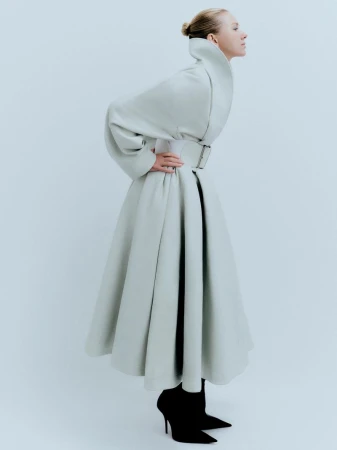 Naomi Watts poses in profile wearing a sculptural flared pale grey Balenciaga coat with a wide belt and black pointed stiletto boots.