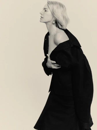 Naomi Watts in profile wearing an off-the-shoulder black Balenciaga jacket against a pale studio backdrop.