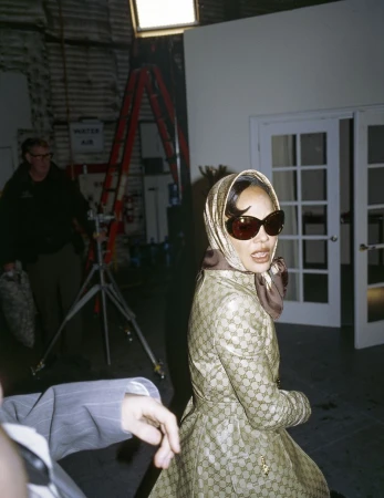 Woman in patterned coat and headscarf, wearing sunglasses, poses in what appears to be a photo studio with equipment in the background.