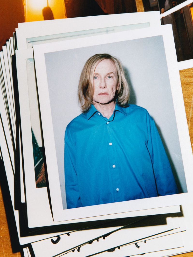 Portrait of an adult with blond hair wearing a blue collared shirt, surrounded by a stack of square photos in a brightly lit indoor setting.