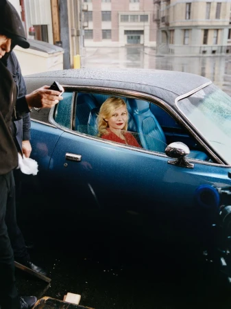 Kirsten Dunst sits inside a classic blue car on set, photographed on a rainy day near a building with crew member visible.