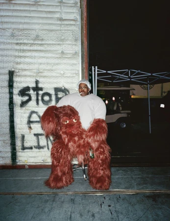 Adult male wearing a costume and standing by an exterior wall with graffiti and an open door at night. Car in the background.