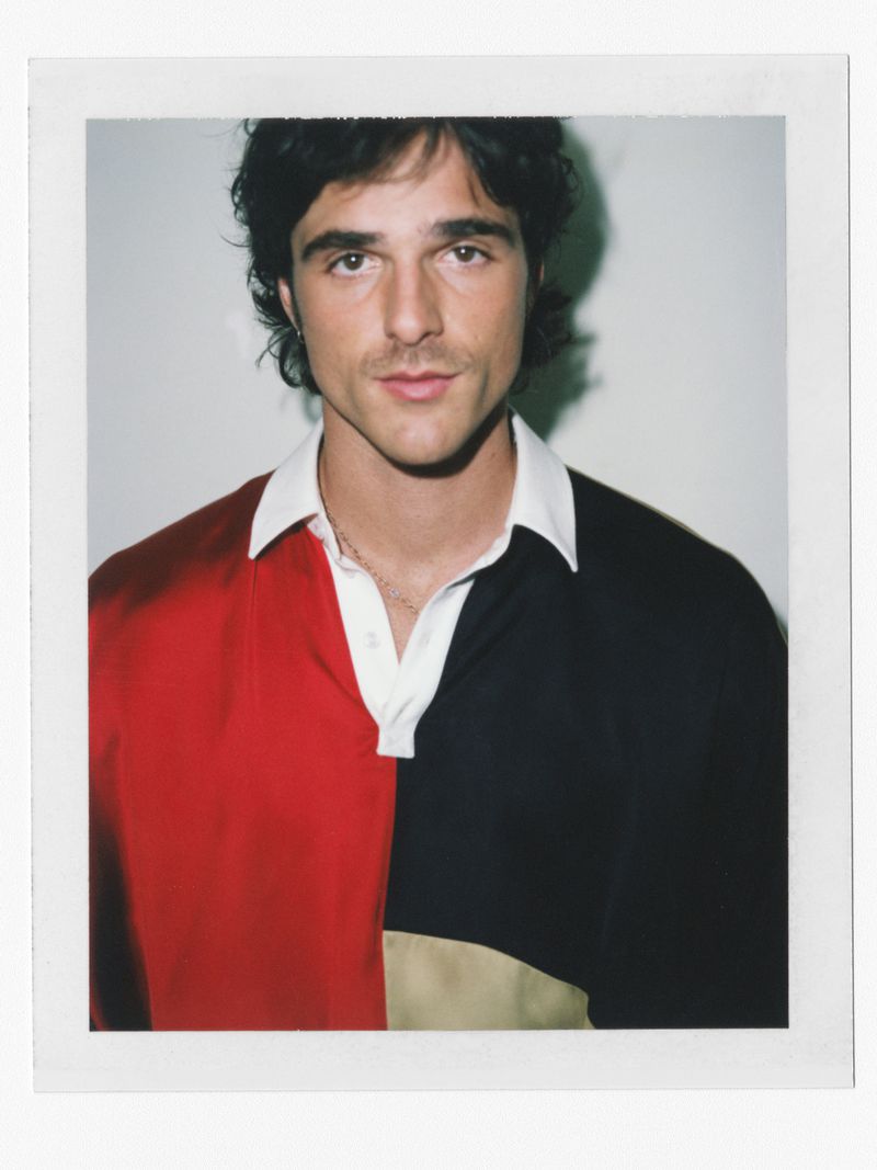 Close-up portrait of a young man with dark hair and a mustache wearing a multi-coloured rugby shirt against a neutral backdrop.