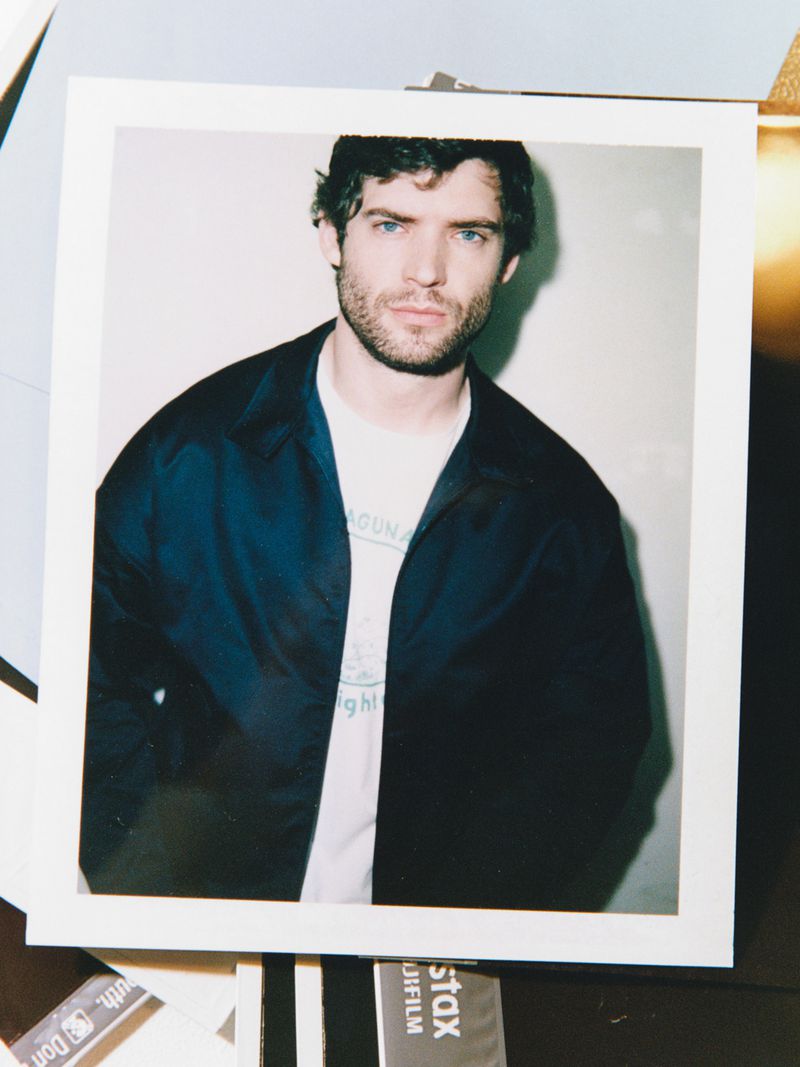 Polaroid photo featuring a dark-haired adult male with blue eyes wearing a jacket and t-shirt in a studio setting, portrait style.