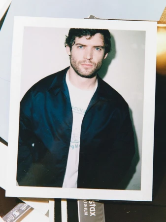 Polaroid photo featuring a dark-haired adult male with blue eyes wearing a jacket and t-shirt in a studio setting, portrait style.