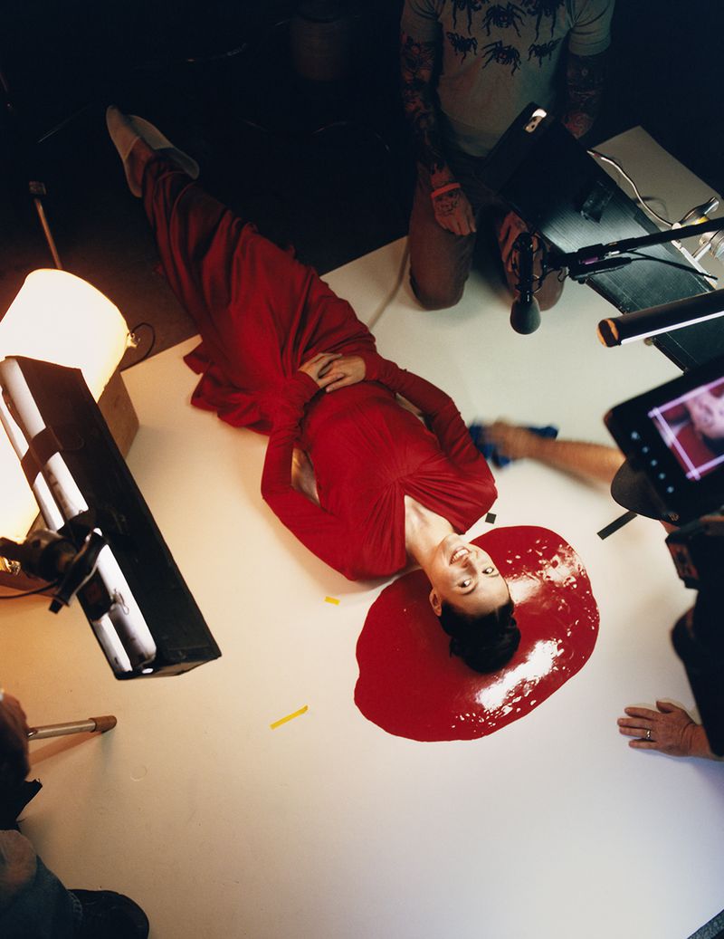 Woman in long red dress poses on white surface surrounded by filming equipment, creating a striking, modern, and visually engaging image.
