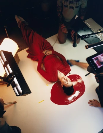 Woman in long red dress poses on white surface surrounded by filming equipment, creating a striking, modern, and visually engaging image.