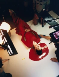 Woman in a long red dress lying on a white studio seamless floor with her head in a pool of red liquid during a photoshoot production.