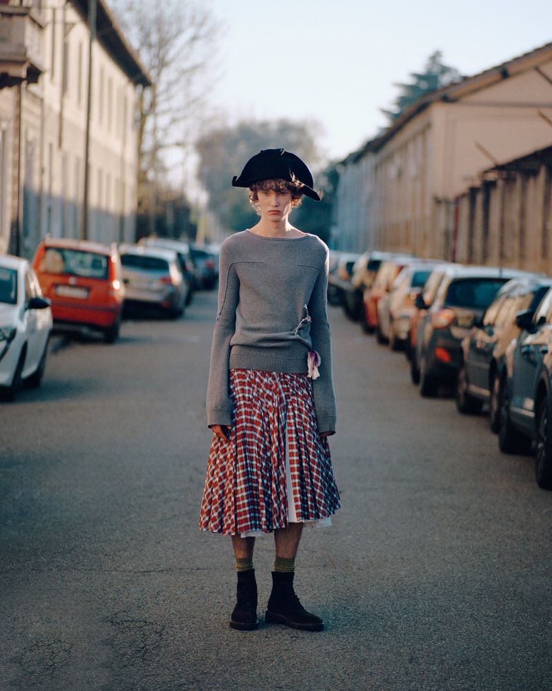 Person in a gray sweater, plaid skirt, and tricorn hat stands on a street lined with cars. Buildings and trees in the background.